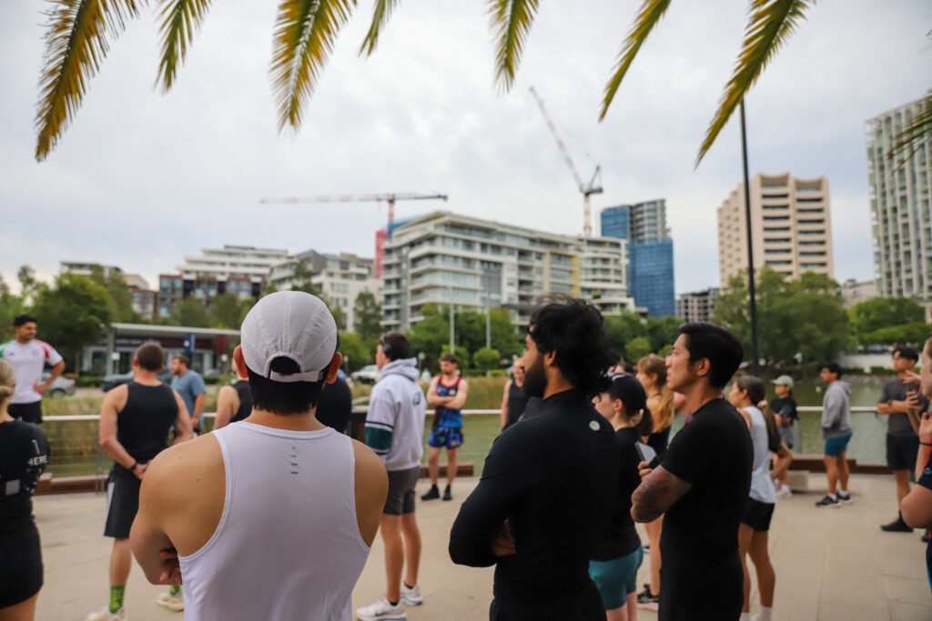 A group of runners standing together on a lakeside path, with palm fronds overhead and apartment buildings behind them