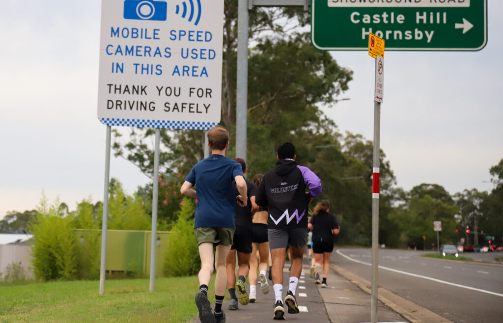 Hills Run Club runners on a footpath near Castle Hill, with a mobile speed camera sign beside the road