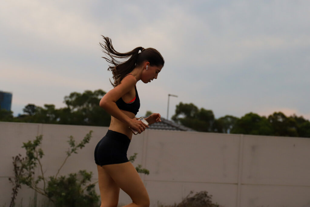 Woman in black activewear running alone along a concrete wall at dusk, with trees behind her