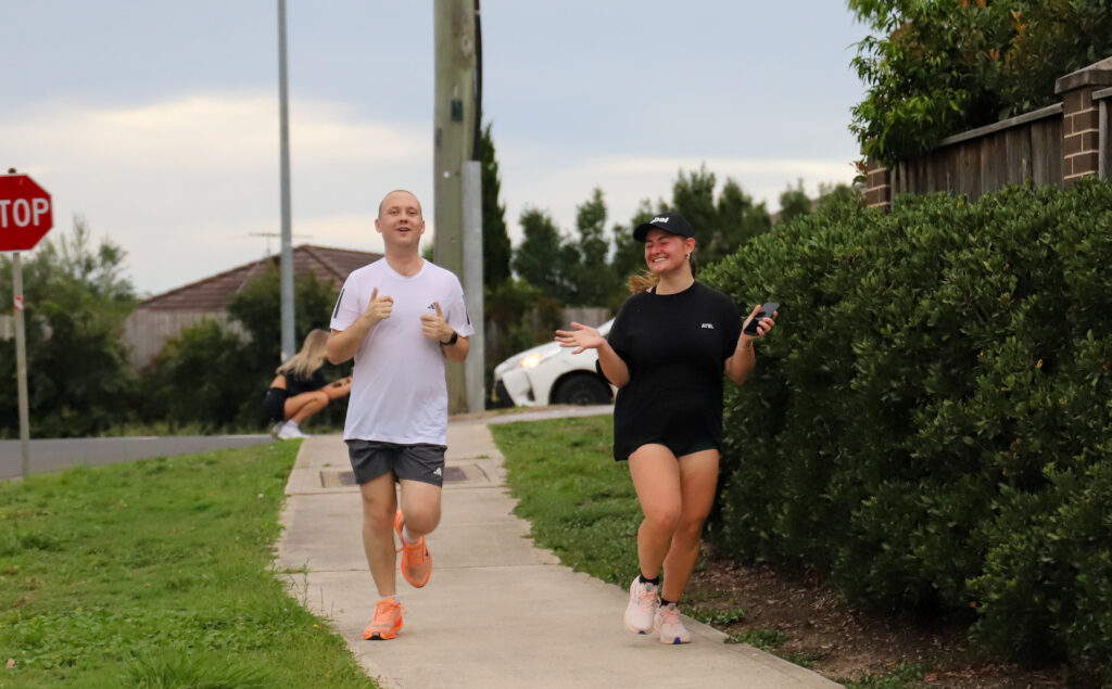Two runners on a footpath, one giving a thumbs up while the other smiles and holds a phone as they run
