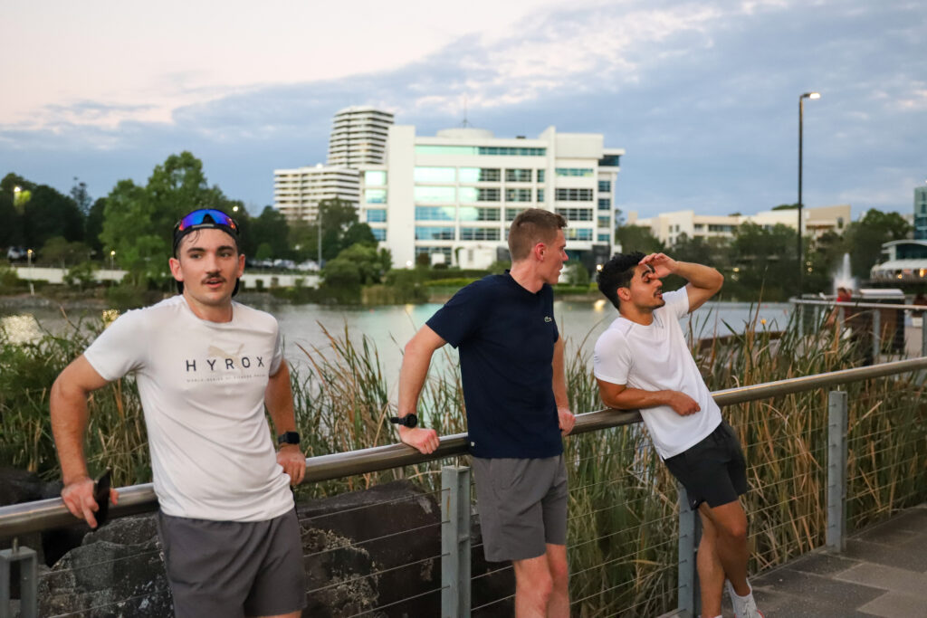 Three runners leaning on a railing beside a lake at dusk, one of them wiping his brow