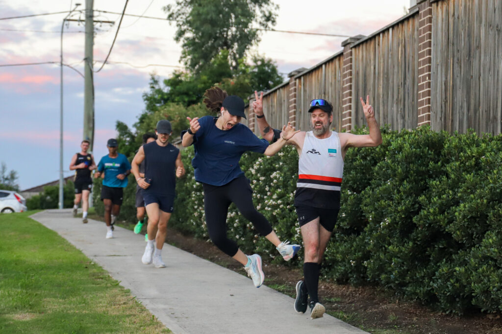 Club runners on a footpath at dusk, one woman jumping mid-stride beside a man giving the peace sign