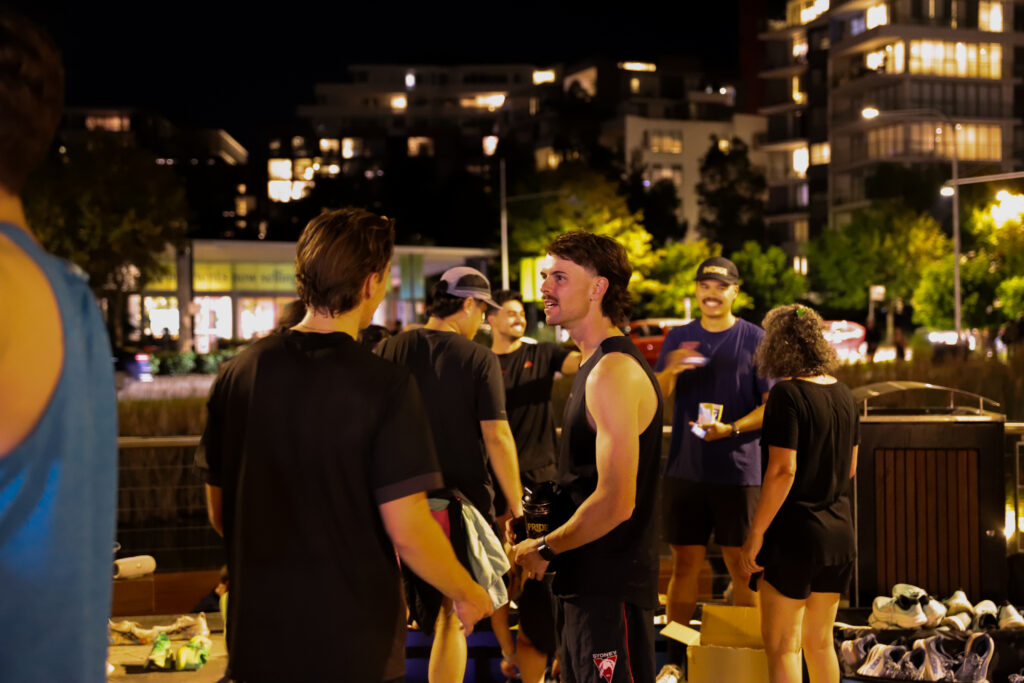 Runners chatting at night on a city footpath beside a row of running shoes laid out on the ground