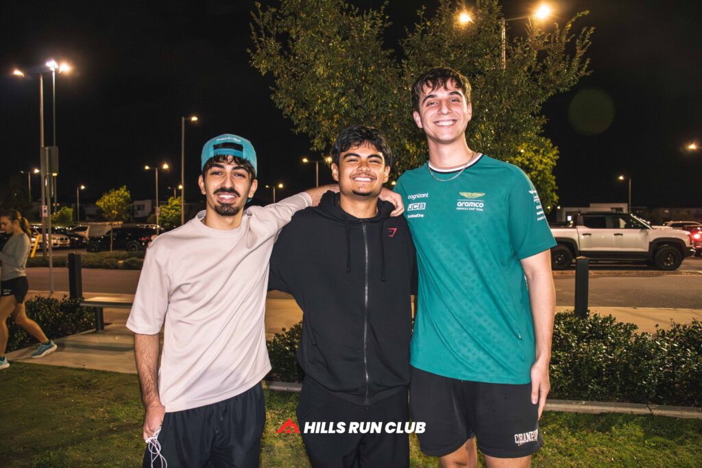 Three runners smiling together under the lights at a Hills Run Club night run