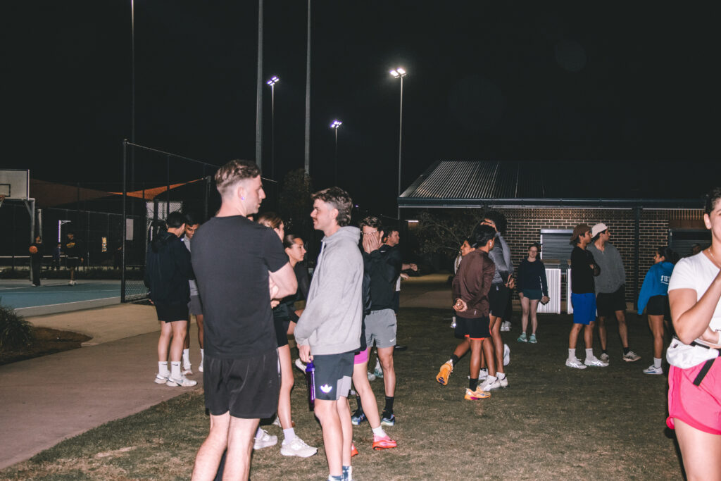 A group of runners standing and chatting on grass under bright lights at night, with a basketball court behind them