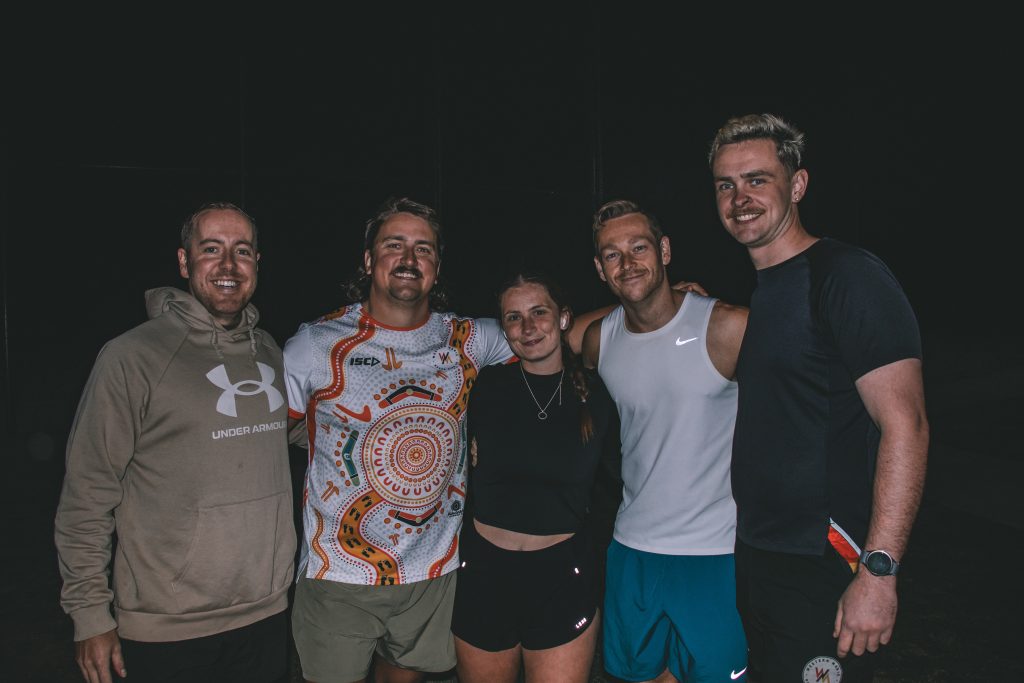 Five members smiling together in the dark after a night run, with a plain black background behind them